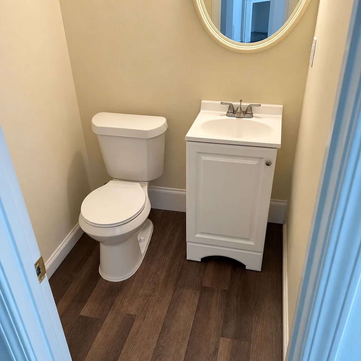 After: the same half bath with plank flooring, a new toilet, a white vanity and trim